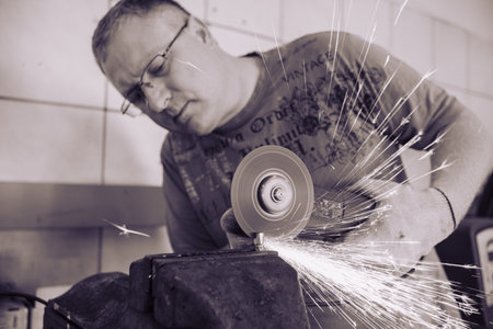 A man works as a grinder in a workshop. A master in glasses grinds a part clamped in a vice with a grinder. Sparks from the metal fly around like a bright fountain.の写真素材