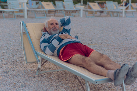 An old man is lying on a sun lounger on the beach. An elderly gray-haired tourist with a beard in red shorts is enjoying himself on the beach by the sea. Grandfather alone on the sea beachの写真素材
