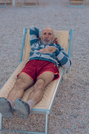 An old man is lying on a sun lounger on the beach. An elderly gray-haired tourist with a beard in red shorts is enjoying himself on the beach by the sea. Grandfather alone on the sea beachの写真素材
