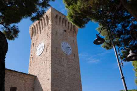 An ancient stone clock tower against a blue sky. Pine trees and a street lamp flank the ancient city...