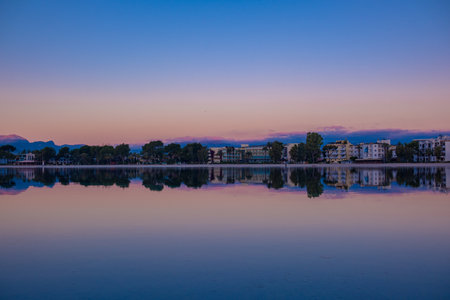 A beautiful view of the city across the mirror surface of the lake at dawn. The city of Port d'Alcudia in Mallorca at dawn. Beautiful pink-blue gradient in the sky and the reflection of the city in the calm water of the lake.の写真素材