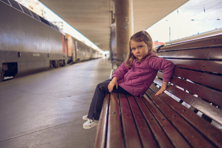 A little girl in a red jacket and jeans sits on a bench at a railway station. A beautiful girl on the platform near a standing train.の写真素材