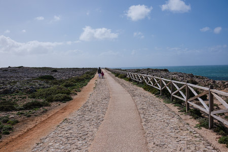 A mother and child walk along a path along the ocean. Mom and daughter in jackets walk along the road by the ocean in sunny and windy weather.の写真素材