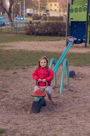 A beautiful little girl plays alone on the playground. The girl is bored alone and has no one to ride with on the teeter-totter or seesaw and she sits on one side of the swing.の写真素材