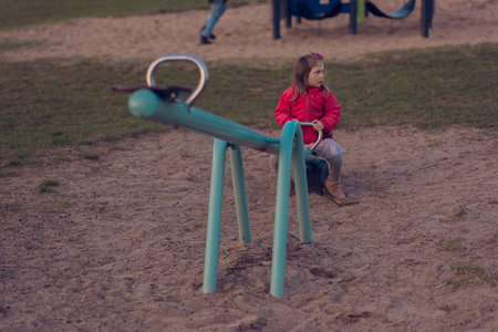 A beautiful little girl plays alone on the playground. The girl is bored alone and has no one to ride with on the teeter-totter or seesaw and she sits on one side of the swing.の写真素材