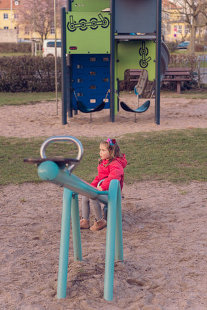 A beautiful little girl plays alone on the playground. The girl is bored alone and has no one to ride with on the teeter-totter or seesaw and she sits on one side of the swing.の写真素材