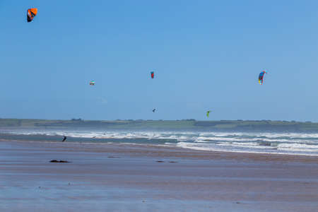 Ireland beach view with kitesurfing athletes. Kitesurfers rush at high speed across the surface of the sea for kites flying in the cloudless sky. Summer sunny day.のeditorial素材