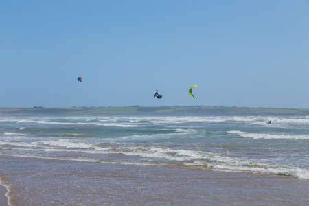 Ireland beach view with kitesurfing athletes. Kitesurfers rush at high speed across the surface of the sea for kites flying in the cloudless sky. Summer sunny day.のeditorial素材