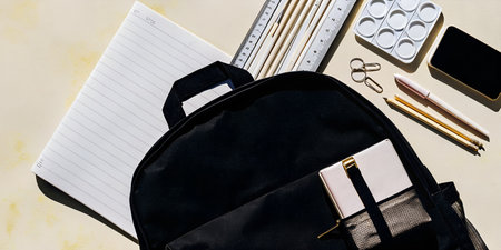 A black backpack filled with school supplies, including notebooks, pencils, and art materials, laid out on a textured light backgroundの素材