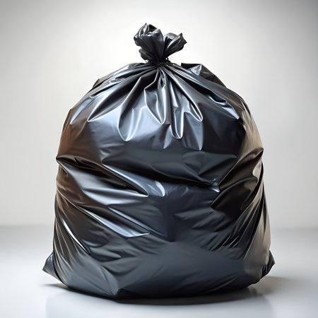 A full black garbage bag tied at the top, standing on a reflective surface with a light gray backgroundの素材
