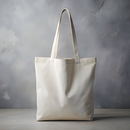 A plain, offwhite canvas tote bag with long handles, set against a textured gray wallの素材