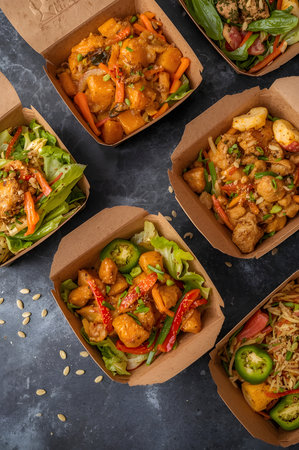 Overhead view of various takeout food boxes filled with delicious asianinspired dishes, arranged on a dark textured surface with scattered seedsの素材