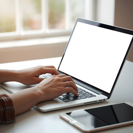 Hands typing on a laptop with a blank screen next to a tablet on a desk by a window with natural lightの素材
