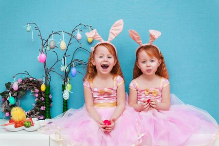 two beautiful girls dressed in pink rabbit ears and lush pink dresses, sitting in their hands, painted chicken eggs in anticipation of the Easter holidayの写真素材