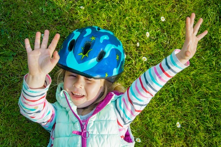girl of three years with a cheerful mood in a protective helmet on her head lies on the green grass with her hands upの写真素材