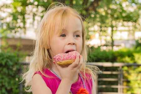 little girl of five years eats beautiful pink donuts outdoors in parkの写真素材