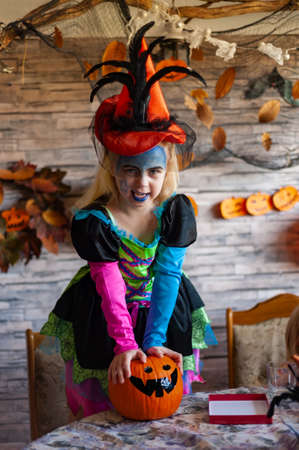 little witch, dressed up girl in a beautiful witch costume holds an orange pumpkin and makes a funny faceの写真素材