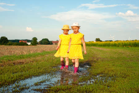 little twin girls in summer yellow dresses and pink boots run through puddles in suburbの写真素材