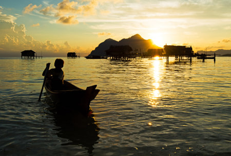 Silhouette of bajau laut young boy paddling a boat during sunrise at the shore of Maiga Island in...