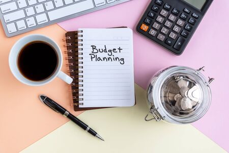 Budget Planning text on note pad with cup of coffee, calculator, computer keyboard, coin jar and pen on colorful pastel colored backgroundの写真素材