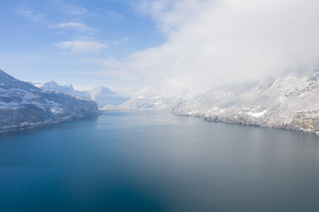 Amazing flight over a beautiful lake in the middle of Switzerland. The cliffs are full of snow and there are many clouds in the sky.の写真素材
