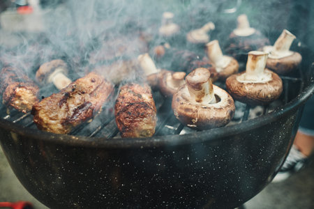 Traditional Romanian mici skinless minced meat rolls and fresh mushrooms grilling over hot coals on a backyard barbecue with thick smoke rising. Romania.