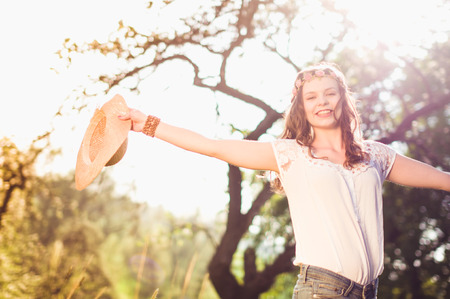 A pretty young woman standing in the back light on a meadow and looking happy at the camera, she is holding a straw hat in her handの写真素材