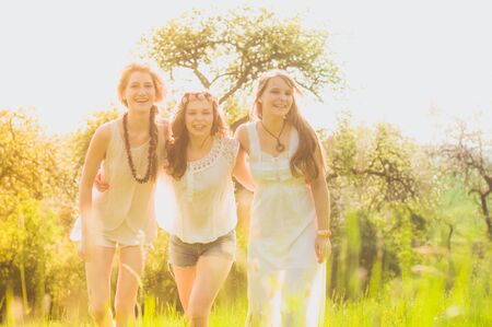 Three young girlfriends in white dresses are omitted arm in arm and laughing at the camera in a meadow in the evening light on a meadow at the edge of the forestの写真素材