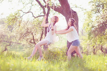 Two young women wear long dresses Wesse in evening light on a meadow. One sits on the swing un the other pushes her to laugh.の写真素材