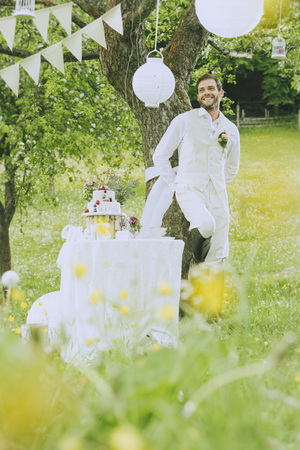 The groom wears a beige vest and is leaning against a tree in the middle of the romantic wedding decoration and smiles.の写真素材