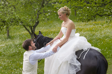 Wedding in the countryside. The bride in her white wedding dress sits on a dark horse and her bridegroom helps her dismount when the horseの写真素材