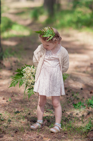 little girl in dress stands in a pine forest and keeps behind a beautiful bouquet of wildflowersの写真素材