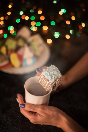 Female's hands with cup of tea and Christmas cookies house on dark color bokeh lights backgroundの写真素材