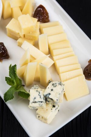 Cheese plate on a dark background table. Many kinds of cheese with sauce and greens on a white plate, closeup shotの写真素材