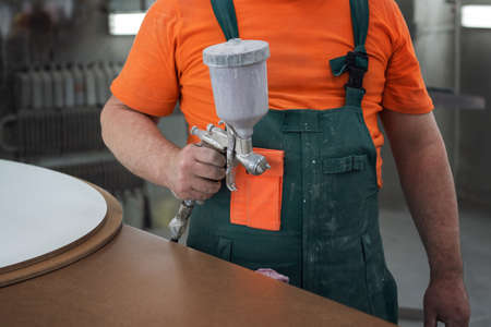 Wooden furniture production. Carpenter painting the surface of the wooden facade with a spray gun
