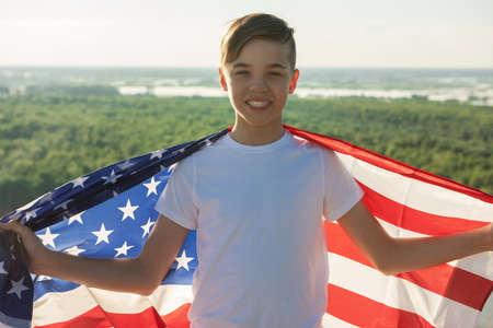 Blonde boy waving national USA flag outdoors over blue sky at the river bankの写真素材