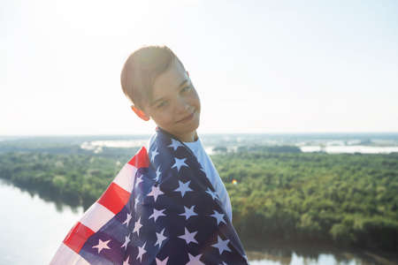 Blonde boy waving national USA flag outdoors over blue sky at the river bankの写真素材