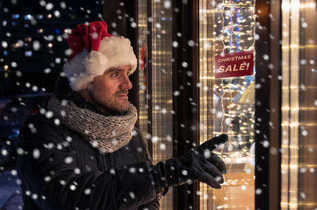 Man in Santas hat looking and dreaming in illuminated shop windowの写真素材
