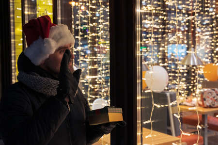 Man in Santas hat looking and dreaming in illuminated shop windowの写真素材