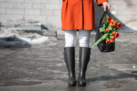 Closeup of woman in a orange coat and rubber boots walking down the street with a bouquet of tulips in spring cityの写真素材