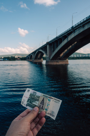Hand with 10 rubles bill on background of Communal bridge in Krasnoyarsk through the Yenisei River in Russia, Krasnoyarsk.の写真素材
