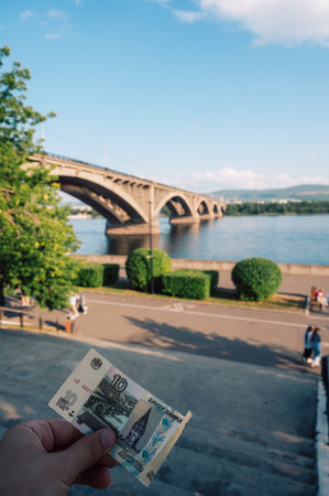 Hand with 10 rubles bill on background of Communal bridge in Krasnoyarsk through the Yenisei River...