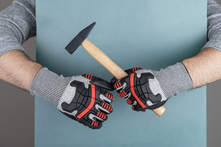 Man's arm in protective gloves of construction worker or building contractor holding hammer on blue paper backgroundの写真素材