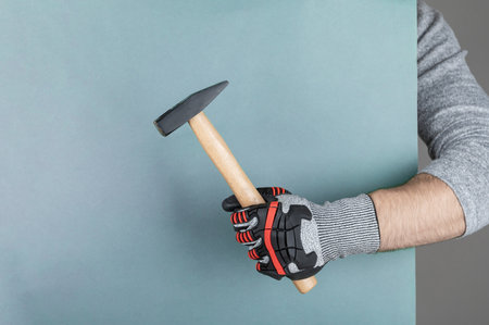 Close-up of a hand wearing a safety glove gripping a hammer, set against a plain background,...