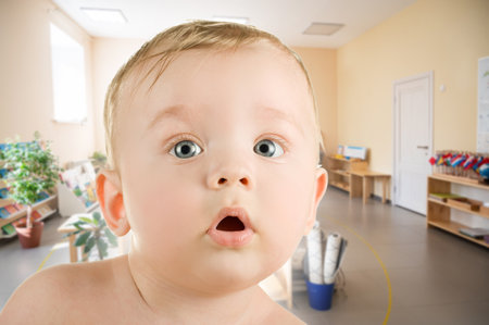 Close-up of a baby with a pacifier in a brightly lit classroom or daycare environment, surrounded by educational materials.の写真素材