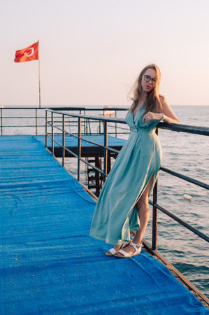 A woman in a flowing green dress poses on a pier by the sea at sunset, with a Turkey flag waving in the background.の写真素材