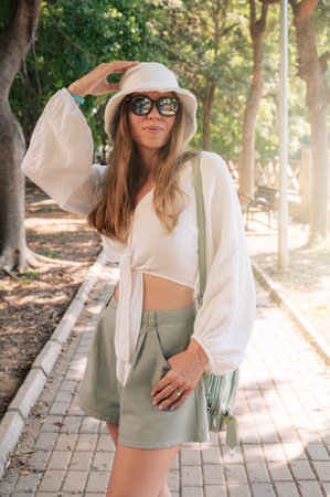 A fashionable woman wearing a white hat, sunglasses, and light summer attire walks along a sunlit park pathway.の写真素材