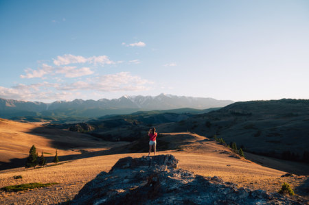 A woman in casual clothing taking photo on her smartphone on a rocky peak, gazing at scenic mountains and a vast valley under golden sunlight.の写真素材