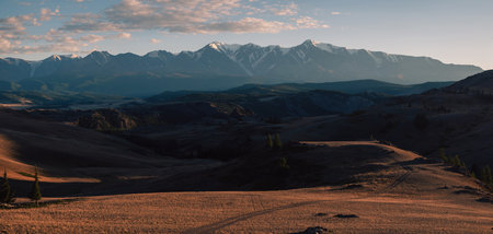 Sunset over a rugged mountain landscape, casting warm golden light across hills, trees, and rocky terrain. Altai Mountainsの写真素材