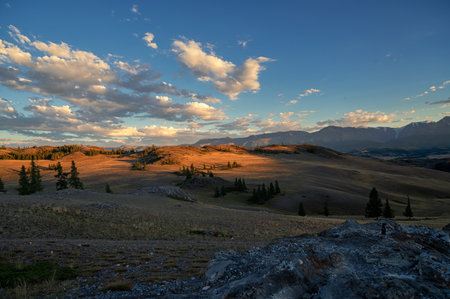 A breathtaking sunset over a rugged mountain landscape, casting warm golden light across hills, trees, and rocky terrain. Altai Mountainsの写真素材